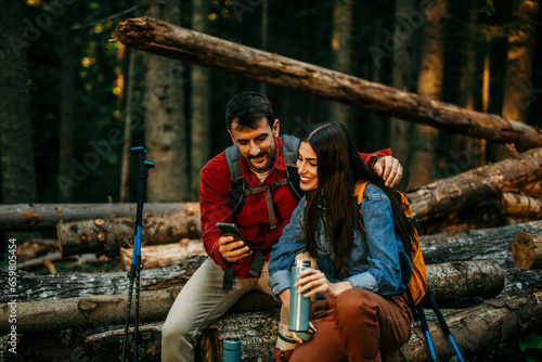Konstfotografi A mixed couple sits together on a sturdy log, radiating a sense of accomplishment and mutual happiness, occasionally checking their phones for any missed notifications