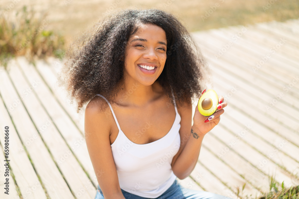 Young African American woman holding an avocado at outdoors smiling a lot Young African American woman holding an avocado at outdoors smiling a lot