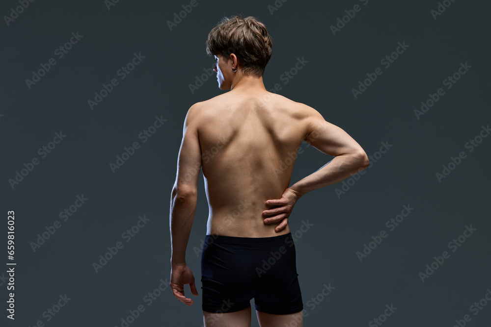 Rear view portrait of young brunette man posing, in underwear holding his muscular, healthy back isolated over dark grey studio background.