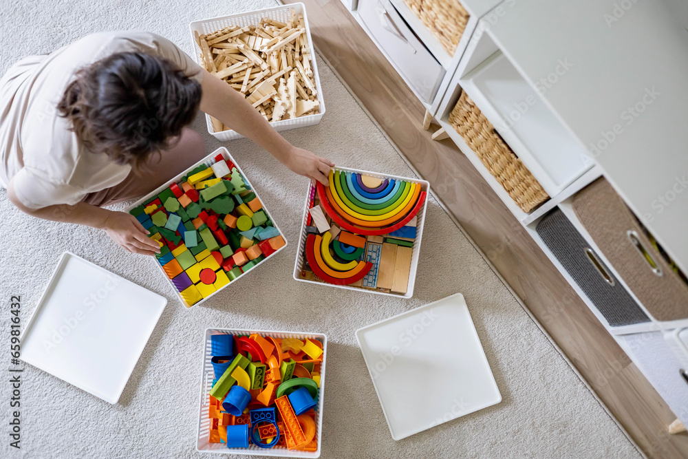 Fototapeta premium Young woman cleaning childish toy sorting wooden Montessori and plastic details box basket top view