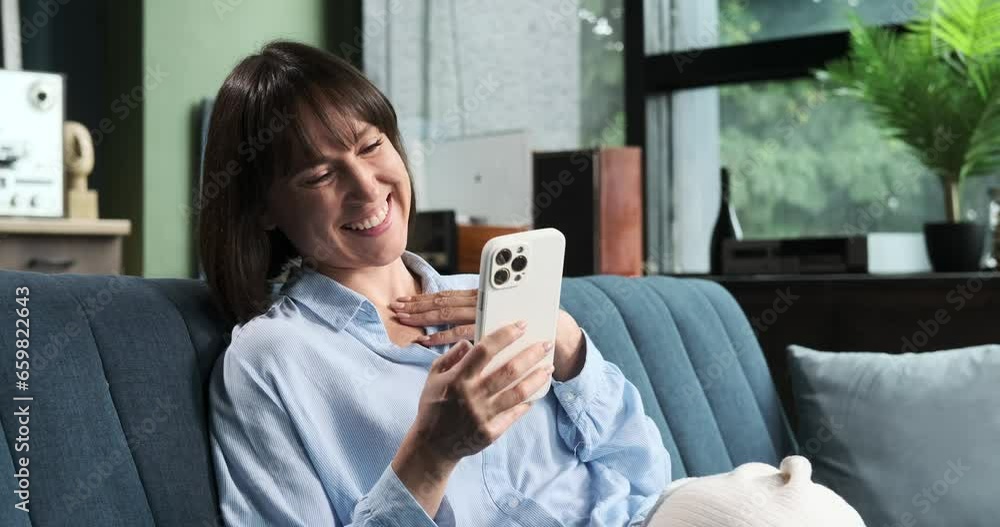 A woman is engrossed in texting on phone while wearing a warm smile in the living room. Her positive and cheerful mood is evident in this cozy and inviting setting. A woman is engrossed in texting on phone while wearing a warm smile in the living room. Her positive and cheerful mood is evident in this cozy and inviting setting.