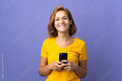 Young Georgian woman isolated on purple background looking at the camera and smiling while using the mobile