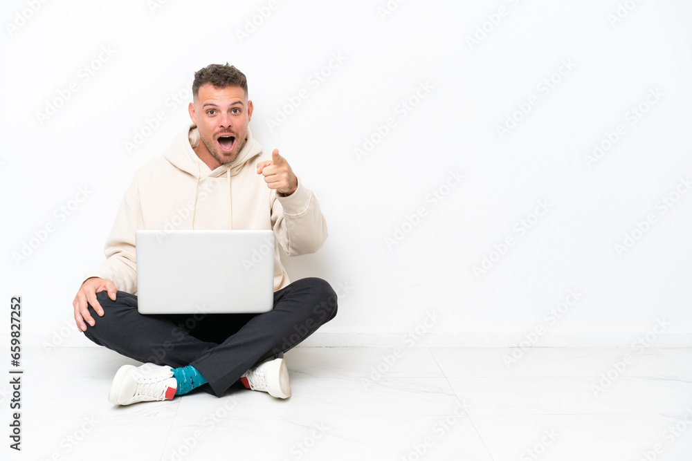 Young caucasian man with a laptop sitting on the floor isolated on white background surprised and pointing front Young caucasian man with a laptop sitting on the floor isolated on white background surprised and pointing front