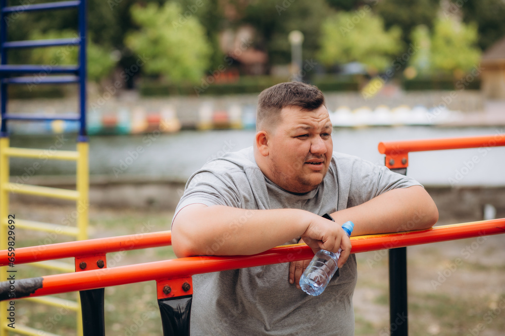 Fototapeta premium fat man eating a burger and drinking water after exercising in the park