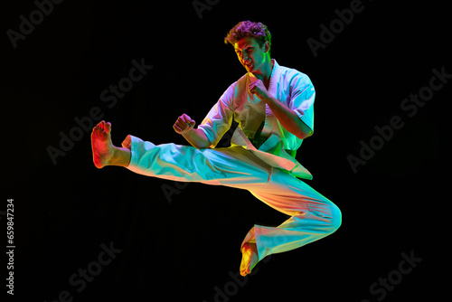 Young sportive man, karateka in white kimono training over black studio background in neon lights. Man standing in karate side kick pose. Concept of martial arts, combat sport, energy, strength. Ad