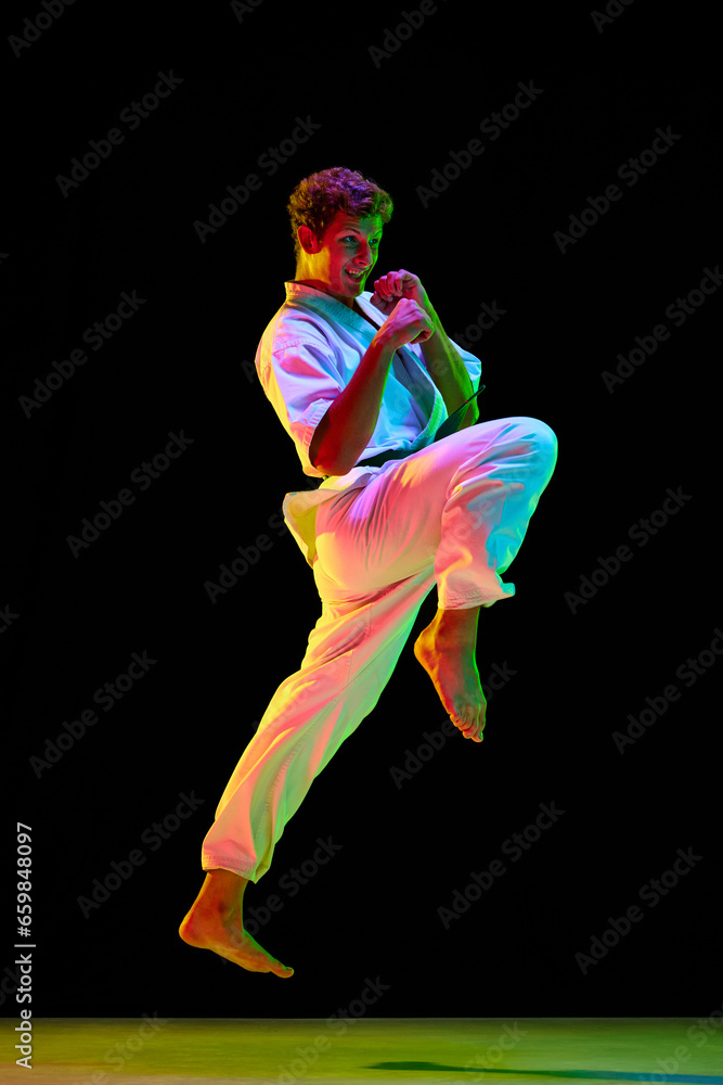 Knee kick. Young man in white kimono practicing karate punches, kicks over black studio background in neon lights. Concept of martial arts, combat sport, energy, strength, health. Ad