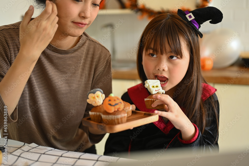 Cute little asian girl dressed as a witch eating colorful Halloween cupcakes with mother in kitchen. People, holiday and festival
