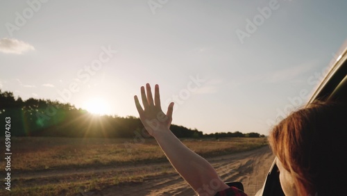 Woman holds hand out of window riding car across countryside at bright sunset