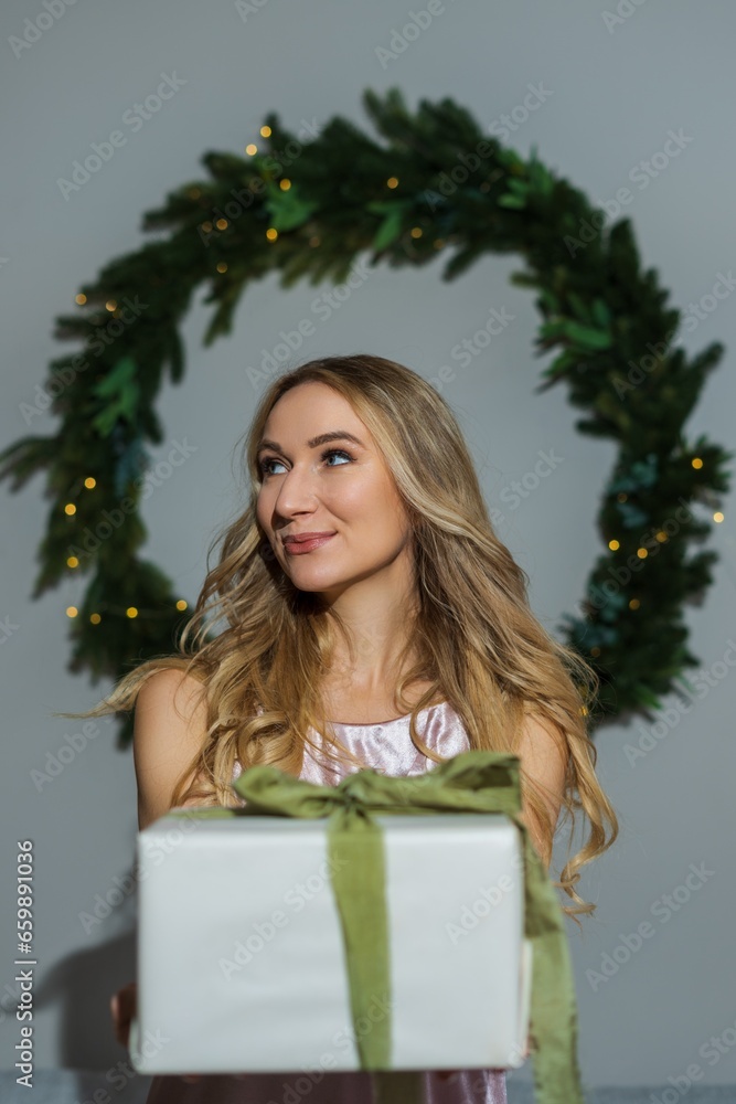 A young well-groomed woman with blond wavy hair and makeup is dressed in a pink dress holding a gift against the background of a New Year's wreath A young well-groomed woman with blond wavy hair and makeup is dressed in a pink dress holding a gift against the background of a New Year's wreath