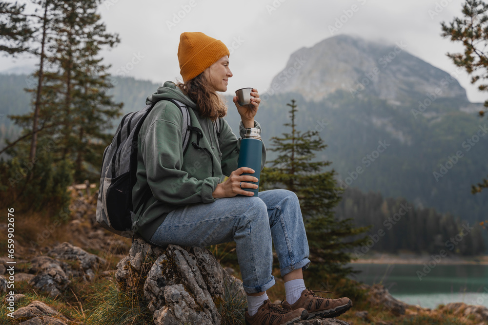 Young cheerful woman traveler with a backpack and thermos on the shore of a lake in the mountains. Solo travel concept - obrazy, fototapety, plakaty 