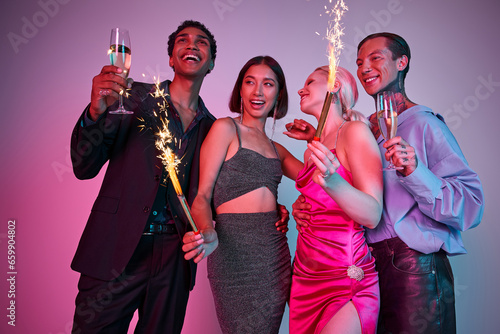 New Year celebration, joyful four multiethnic friends holding sparklers and champagne on purple pink