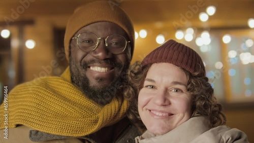 Portrait medium close-up shot of ethnically diverse family couple wearing warm clothes standing against cottage, looking at camera photo