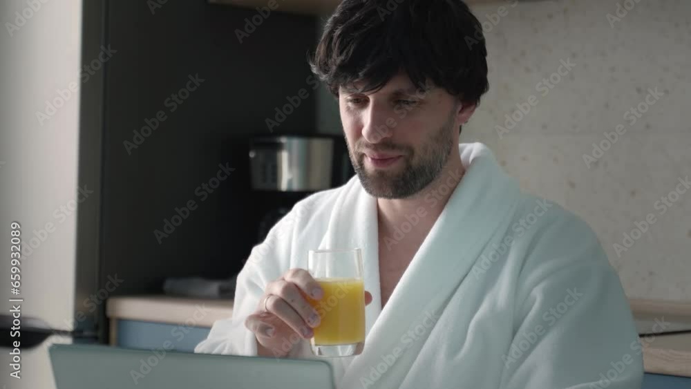 A black-haired man in a bathrobe drinks orange juice, works on a laptop in the kitchen. A black-haired man in a bathrobe drinks orange juice, works on a laptop in the kitchen.