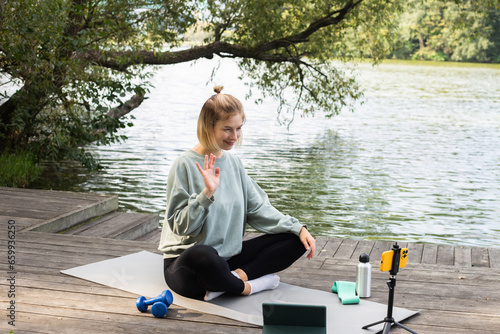 Woman doing exercises in nature near the lake. Yoga mat, bottle, weights, tripod with live camera. A person leads an online fitness class and waves his hand at the camera