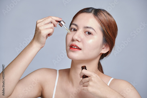 Asian woman applying serum on her face with pleasure. Black head girl holding dropper with skin care product for healthy and glowing skin tone, isolated background