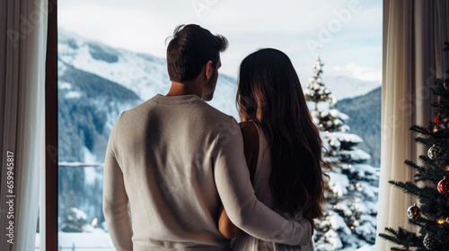 A man hugs a woman at the window of a New Year's decoration hotel room with snowy mountains view.
