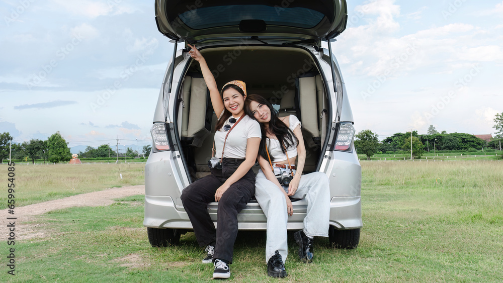 Two young Asian women in white shirts and jeans sit in the back of their new car, lounging on the field. holidays and traveling