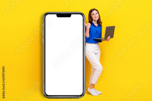 Full length photo of sweet beautiful lady wear blue shirt pointing thumb back modern device empty space isolated yellow color background