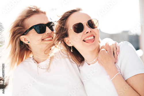 Two young beautiful smiling hipster female in trendy summer white t-shirt and jeans clothes. Carefree women posing in the street. Positive models having fun outdoors. Cheerful and happy. In sunglasses