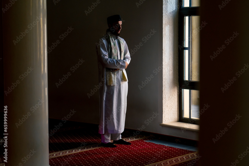 Muslim believer in national clothes prays in the twilight by the window in the mosque kneels down and turns to Allah