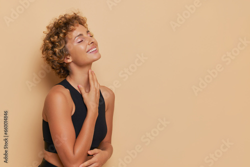 Pretty female model in black sports top standing with hands on her chest against beige background, lifestyle concept, copy space