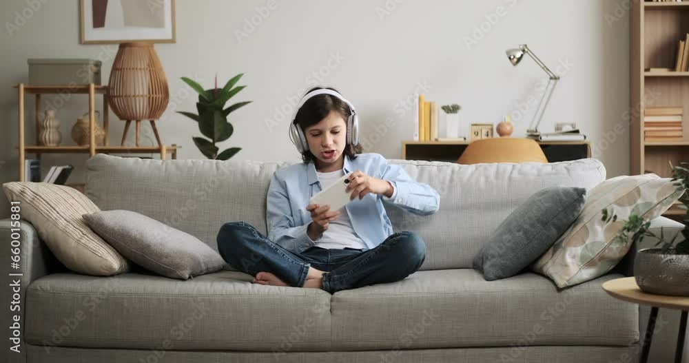 Determined schoolboy sits on a couch, engrossed in a mobile game. His unwavering focus on the screen while playing creates an atmosphere of concentration and dedication, showcasing enthusiasm. Determined schoolboy sits on a couch, engrossed in a mobile game. His unwavering focus on the screen while playing creates an atmosphere of concentration and dedication, showcasing enthusiasm.