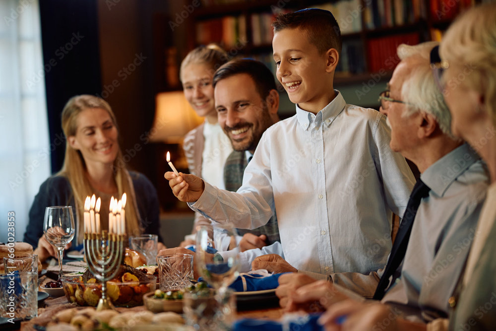 Happy Jewish boy lighting menorah during family meal on Hanukkah.