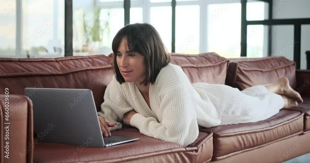 Cheerful Caucasian woman lies on the sofa, a warm smile gracing face as she operates laptop. Her relaxed position and positive expression illustrate a harmonious blend of comfort and productivity. Cheerful Caucasian woman lies on the sofa, a warm smile gracing face as she operates laptop. Her relaxed position and positive expression illustrate a harmonious blend of comfort and productivity.