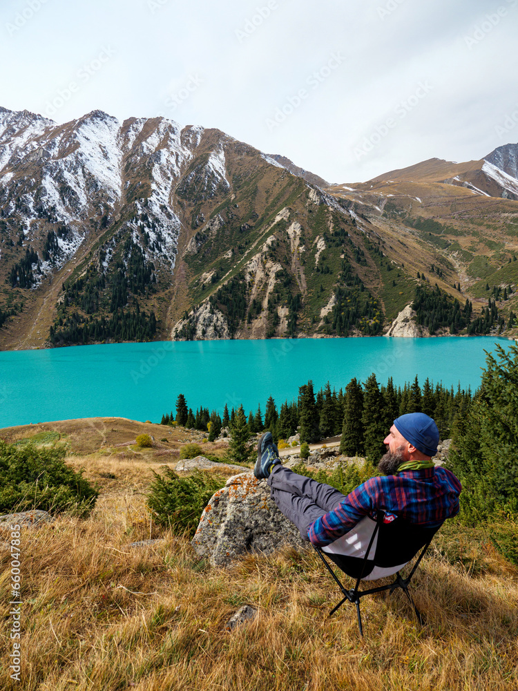A bearded man sitting on a folding chair rests in the mountains on the shore of a beautiful lake. We enjoy a magnificent panoramic view of snow-capped peaks and a turquoise mountain lake A bearded man sitting on a folding chair rests in the mountains on the shore of a beautiful lake. We enjoy a magnificent panoramic view of snow-capped peaks and a turquoise mountain lake