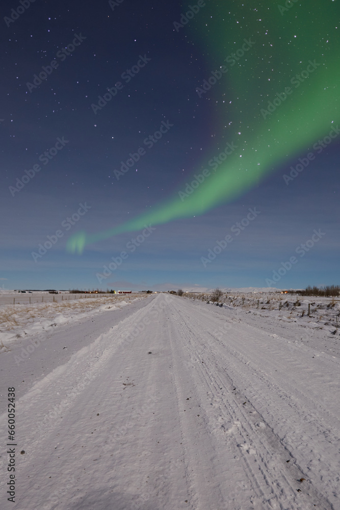 A vivid green aurora borealis dances across a starlit sky, illuminating a snow-covered road and distant mountains.
