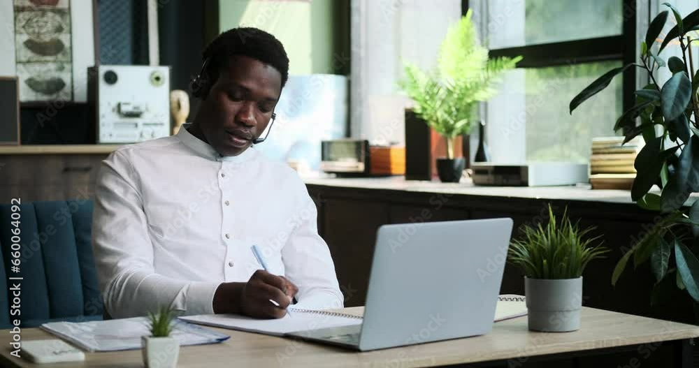 A professional Black manager, equipped with a headset, conducts a business call from the comfort of home. His efficient and focused demeanor reflects his commitment to work responsibilities. A professional Black manager, equipped with a headset, conducts a business call from the comfort of home. His efficient and focused demeanor reflects his commitment to work responsibilities.