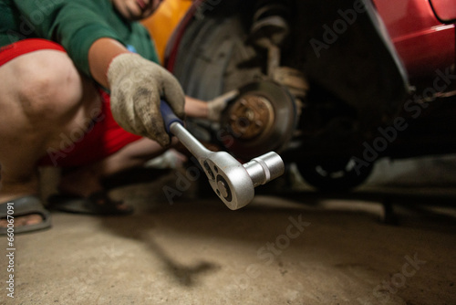 A man repairing a car in a garage holds a ratchet wrench in the cell. A man takes a ratchet wrench to remove rusty bolts from a car wheel hub. Car repair in the garage.