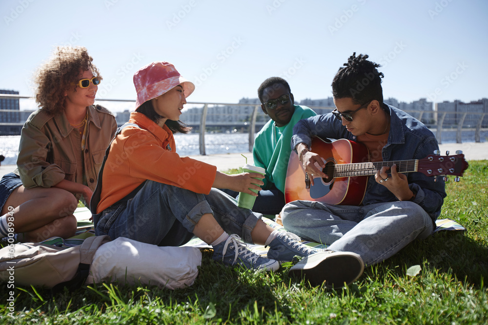 Multiethnic group of friends playing guitar outdoors while enjoying picnic on grass in city