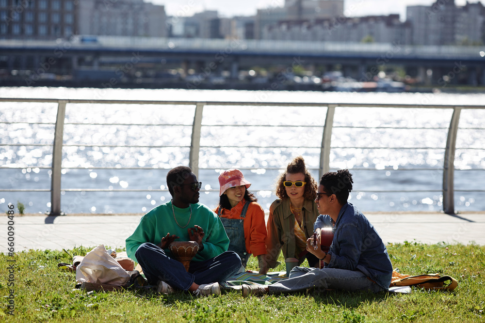 Wide angle view of young people enjoying picnic on grass outdoors with city skyline in background, copy space
