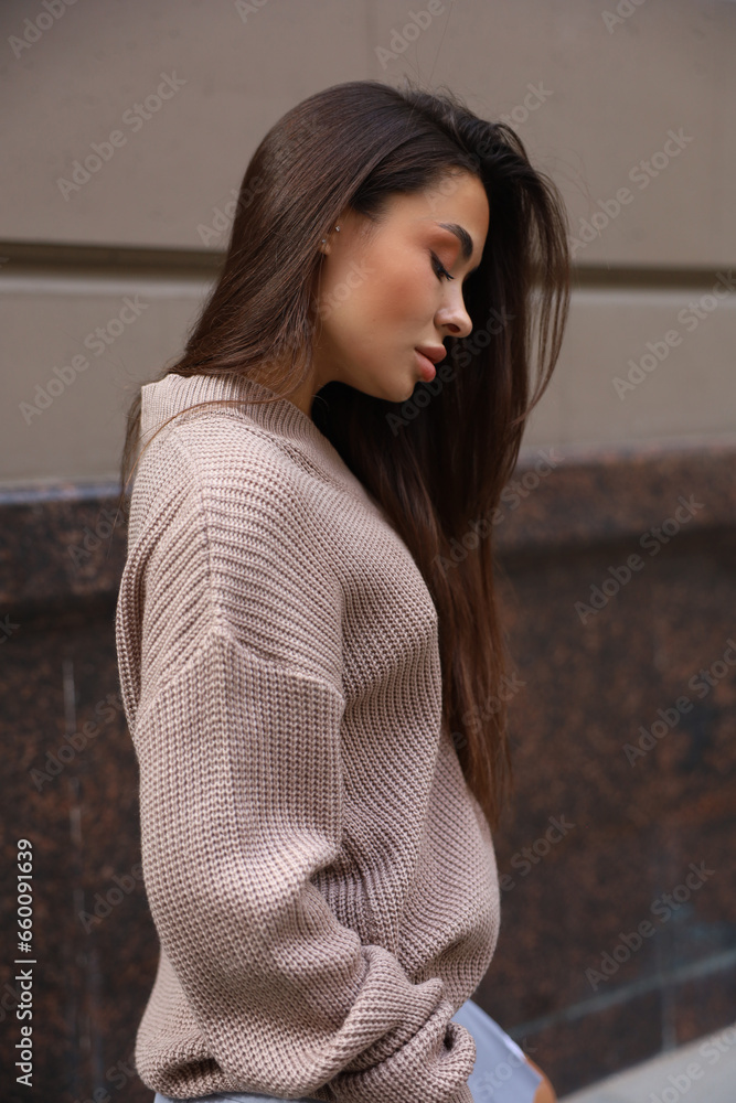 A brunette woman on the street in a brown sweater and a blue skirt next to a beige building. everyday lifestyle. casual A brunette woman on the street in a brown sweater and a blue skirt next to a beige building. everyday lifestyle. casual