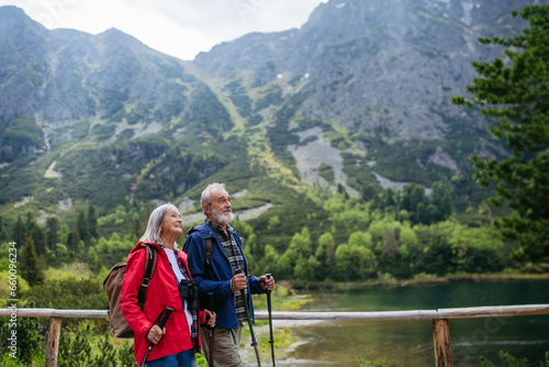 Active elderly couple hiking together in autumn mountains, on senior-friendly trail. Walking by lake, enjoying nature. Senior tourists using trekking poles for stability.