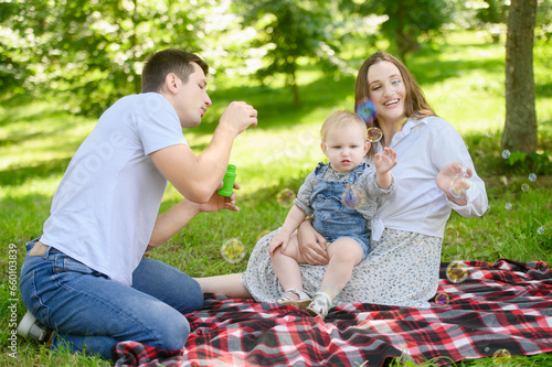 A young cheerful family with a child is having a picnic in the park, dad is blowing soap bubbles.Summer time.Family day © GRON777