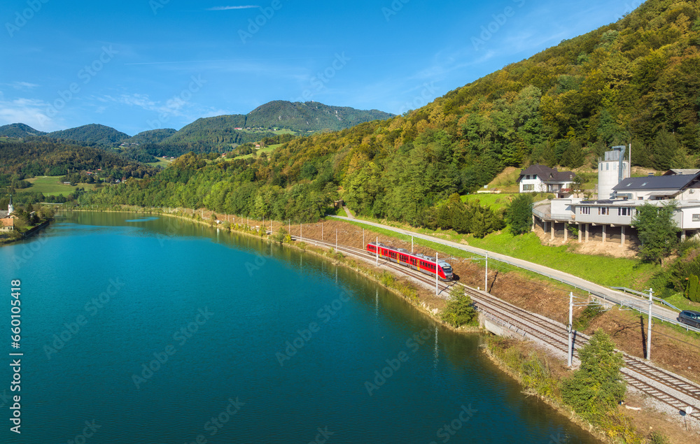 Fototapeta premium Aerial view of red modern high speed train moving near river in alpine mountains at sunset in autumn. Top view of train, rural railroad, lake, road, green trees in fall. Railway station in Slovenia