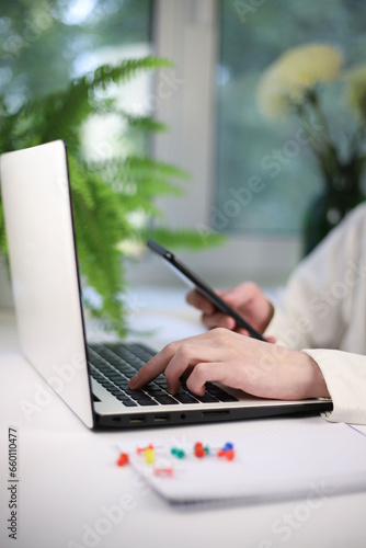 Online shopping concept. There is a white laptop on a white table. next to it is a notepad for notes and a worker’s hands on a keyboard and smartphone