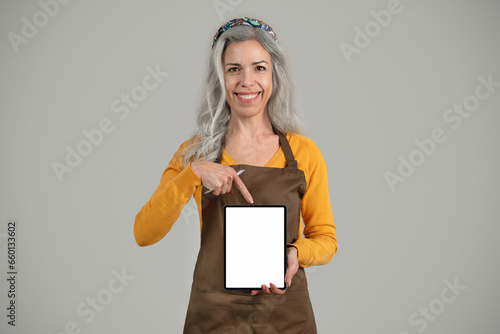 Smiling senior european woman gray hair framer in apron point finger at banner with free space photo
