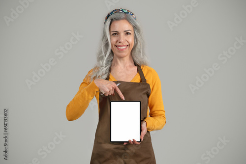 Foto Smiling senior european woman gray hair framer in apron point finger at banner w