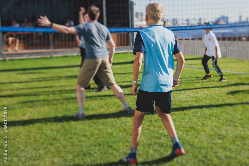 Fototapeta premium Volleyball game, junior teenage school team of kids play volleyball, players on the outdoor playground with net and green lawn grass court, sports children team during the game, summer sunny