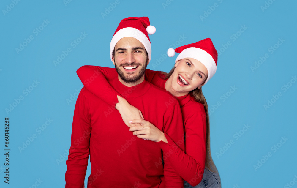 Happy Holidays. Cheerful Young Couple In Santa Hats Posing On Blue Background Happy Holidays. Cheerful Young Couple In Santa Hats Posing On Blue Background