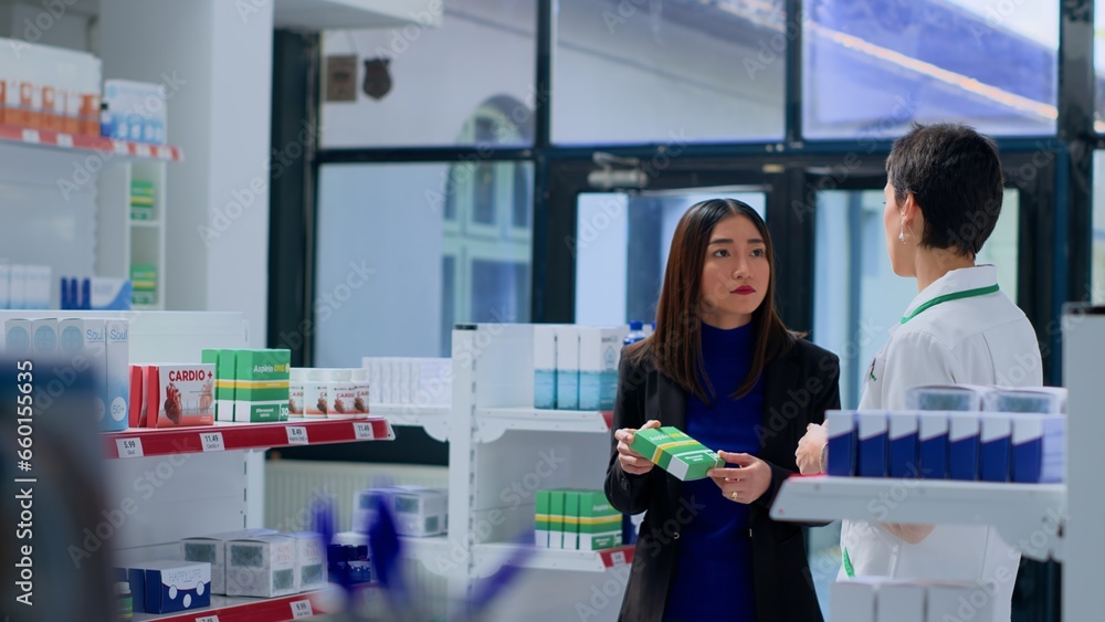 Asian woman in dispensary needing prescription drug to fight off virus, asking druggist to help her pick best medicament. Pharmacy technician assisting customer with product sugestion Asian woman in dispensary needing prescription drug to fight off virus, asking druggist to help her pick best medicament. Pharmacy technician assisting customer with product sugestion