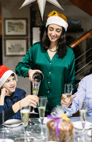 Happy family celebrating christmas vacation at home. Young beautiful woman reunited with her family pours champagne into glass. New year and xmas concept. Vertical photo.