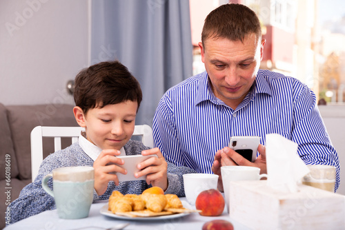 Happy tween boy spending time with his father at home  carried away with mobile gadgets during breakfast