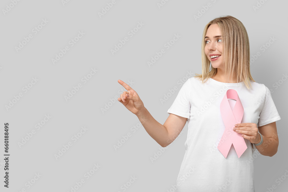 Young woman with pink ribbon pointing at something on grey background. Breast cancer awareness concept Young woman with pink ribbon pointing at something on grey background. Breast cancer awareness concept