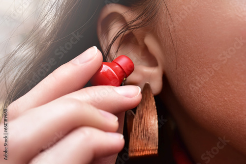 A woman holds a red wireless earphone in her hand, bringing it closer to her ear to listen to music. photo