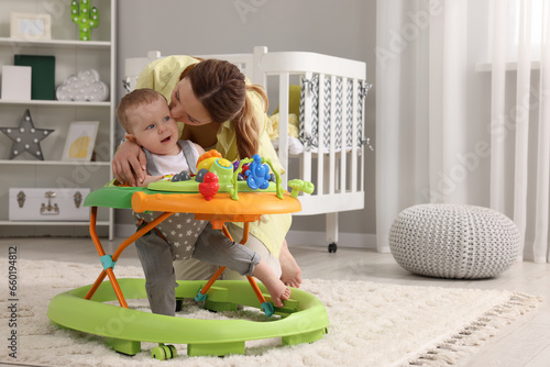 Mother kissing her little son at home. Cute baby making first steps with toy walker, space for text photo