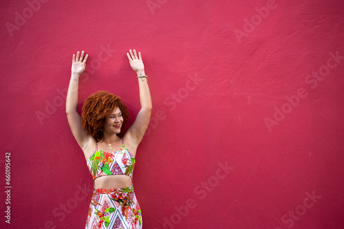 Portrait of a beautiful red-haired woman in a long, colorful dress standing with her arms moving against a red wall.